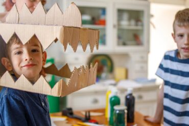 A woman placing a cardboard dinosaur head on a smiling boy beside a cluttered crafting counter. There is another child nearby.