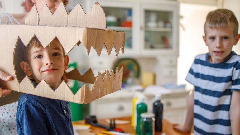 A woman placing a cardboard dinosaur head on a smiling boy beside a cluttered crafting counter. There is another child nearby.