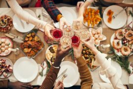A top-down shot of a dinner table and six people extending glasses out to cheers over a spread of food.
