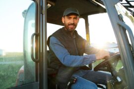 A farmer smiles as he sits in the cab of his tractor with the door open. The sun is visible in the background.