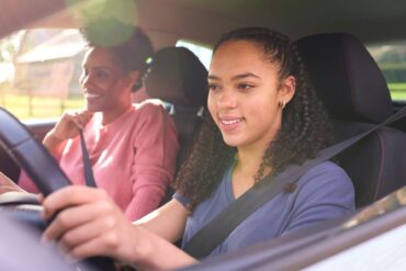 A Black woman in a pink shirt sits in the passenger seat, and a young Black girl drives with the window rolled down.