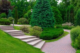 A beautifully landscaped yard contains a brick pathway, evergreen trees, white flowers, and mature bushes.