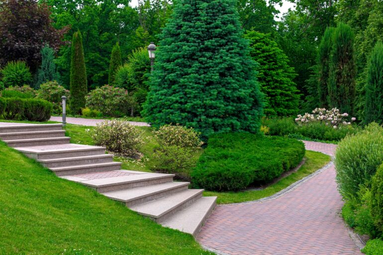 A beautifully landscaped yard contains a brick pathway, evergreen trees, white flowers, and mature bushes.