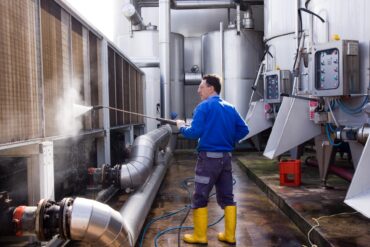 A man wearing a blue shirt and protective gear uses a high-pressure water spray to clean industrial equipment.