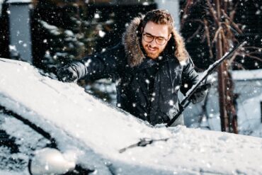 A man scaping snow off of his windshield. He is wearing glasses and a black puffy coat. There is snow in the air.