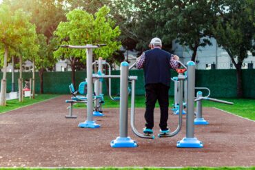 An older person wearing a white hat uses the leg workout equipment in an outdoor public park surrounded by trees.