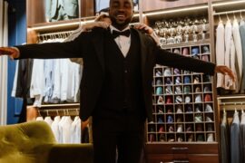 A man smiles as he stands in a tailor shop and wears a tuxedo with his arms stretched out. The tailor adjusts the coat.