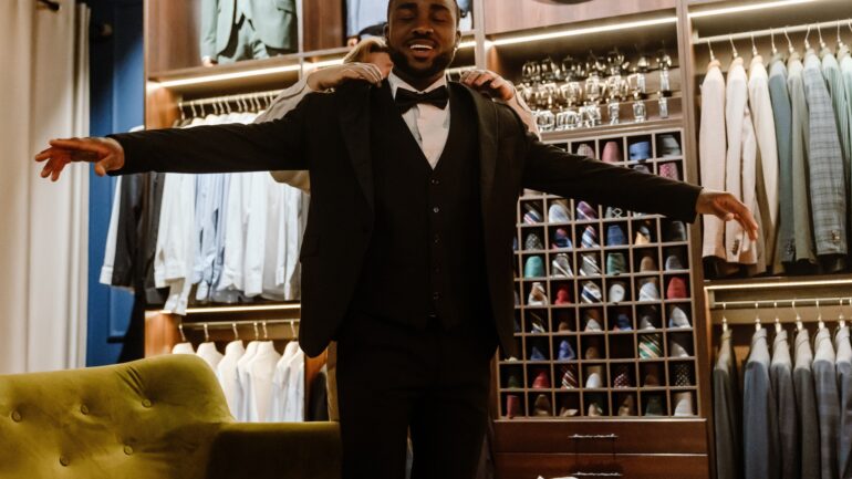 A man smiles as he stands in a tailor shop and wears a tuxedo with his arms stretched out. The tailor adjusts the coat.