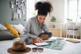 A young woman with curly hair is sitting on her bed and using her tablet and a map indoors at home to plan a trip.