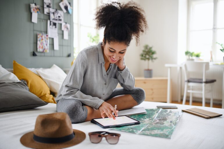 A young woman with curly hair is sitting on her bed and using her tablet and a map indoors at home to plan a trip.