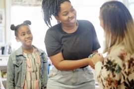 An elementary school teacher warmly greeting the mother of the two students standing behind her.