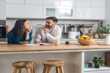 A man and a woman lean on the counter of a kitchen island. They have a pondering look on their faces.