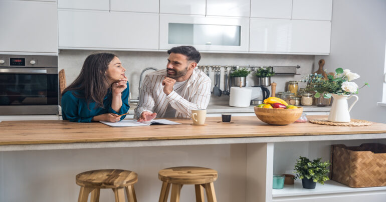A man and a woman lean on the counter of a kitchen island. They have a pondering look on their faces.