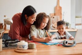 A homeschooling mother standing behind her young daughter and even younger son doing homework at the kitchen table.