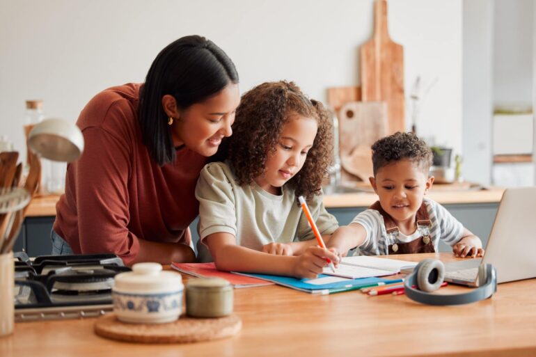 A homeschooling mother standing behind her young daughter and even younger son doing homework at the kitchen table.
