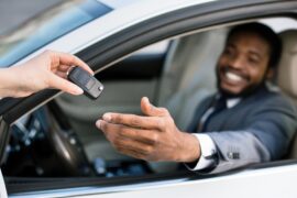 A person is handing a black key fob to another person sitting in the driver's seat of a black car through the open window.