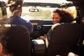 A mom smiles as she sits in a car, looking back at her children in the rear seats. Her husband drives the vehicle.