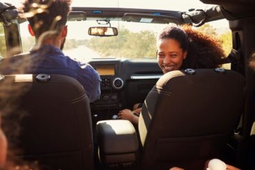 A mom smiles as she sits in a car, looking back at her children in the rear seats. Her husband drives the vehicle.