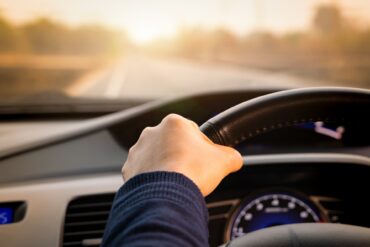 A close-up of a person's hand gripping a vehicle's steering wheel, and the open road visible in the distance.