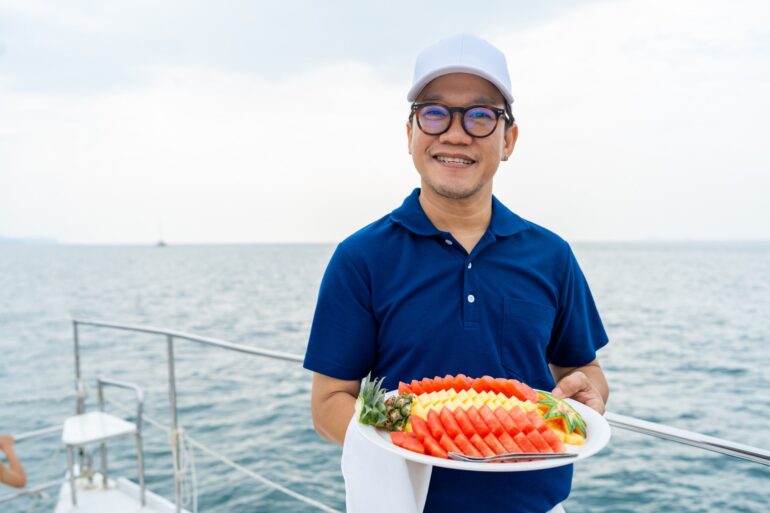 A man wearing a blue polo and a white cap, holding a large tray with fruit on the edge of a boat floating in the ocean.