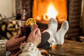 A woman holding a glass of wine, relaxing with her feet up on the table. There is a fire roaring in the background.