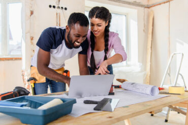 A young couple smiles and looks at a laptop on a workbench in an unfinished room with tools and blueprints strewn about.