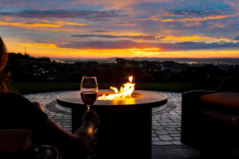 A close-up of the arm of a woman holding a glass of wine next to a large fire pit in a patio while watching a sunset.