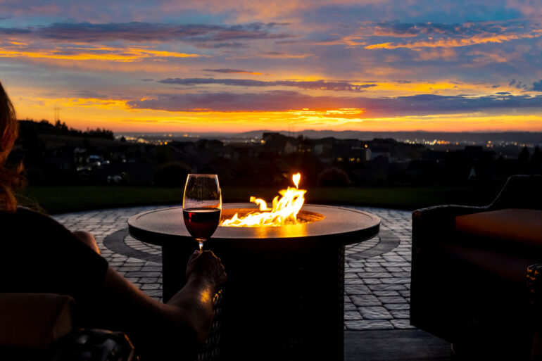 A close-up of the arm of a woman holding a glass of wine next to a large fire pit in a patio while watching a sunset.