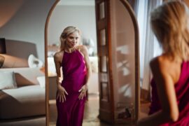 A blond woman with short hair looking in the mirror in her bedroom. She is wearing a long pink dress.