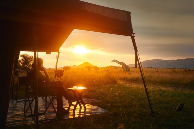 A woman sitting outside a luxury tent in Africa. She is watching the sunset and the field in front of her.