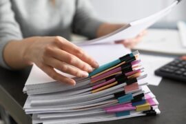 edentials: New Africa, 251700536 Alt text: A close-up of a hand touching a large pile of documents piled on a desk while another hand holds one document up.