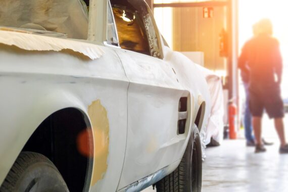 A close-up of the side of a white restoration car parked in the garage. There are people standing in the garage door.