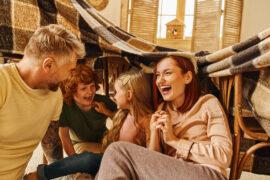 A mom, dad, and their son and daughter laugh together while sitting under a makeshift chair-and-blanket fort.