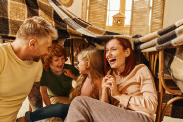 A mom, dad, and their son and daughter laugh together while sitting under a makeshift chair-and-blanket fort.
