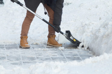 A close-up of a person's legs wearing khakis and brown boots and a black coat while scooping snow shoveling.