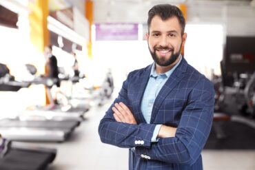 A male gym business owner stands in the foreground of his gym with a blurry view of gym equipment directly behind him.