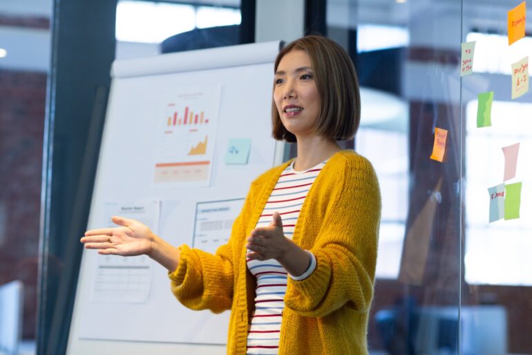 A woman stands in an office and gives a presentation with graphs and other papers behind her. She wears a yellow sweater.