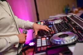 A close-up of a DJ's hands on a mixing console with glowing buttons, holding a microphone, under vibrant purple lighting.