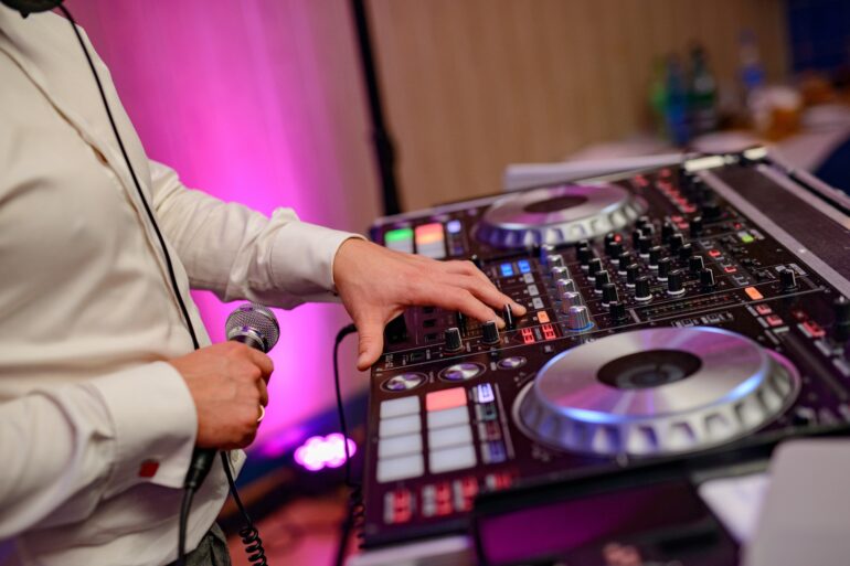A close-up of a DJ's hands on a mixing console with glowing buttons, holding a microphone, under vibrant purple lighting.