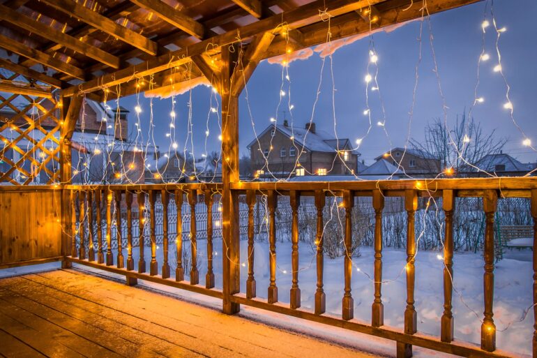 A home's covered wooden deck at dusk in the winter. The floor is dusted with snow, and string lights hang from above.