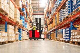 A person is operating a forklift in the middle of a warehouse aisle. There are boxes stacked up on pallet racks.