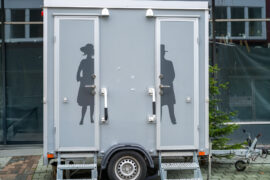 An outdoor restroom trailer with a stall designated to each gender. The trailer is gray with labeled doors.