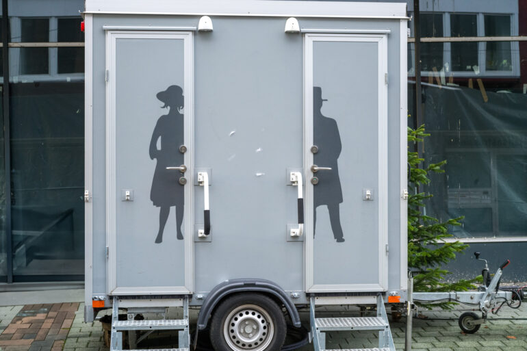 An outdoor restroom trailer with a stall designated to each gender. The trailer is gray with labeled doors.