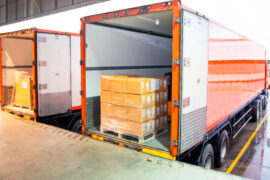 Two semi-trucks parked at a loading dock with their trailer doors open, each holding pallets with stretch-wrapped boxes.