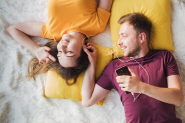 A man and woman on a blanket with pillows, sharing a pair of earbuds connected to a phone the man is holding.