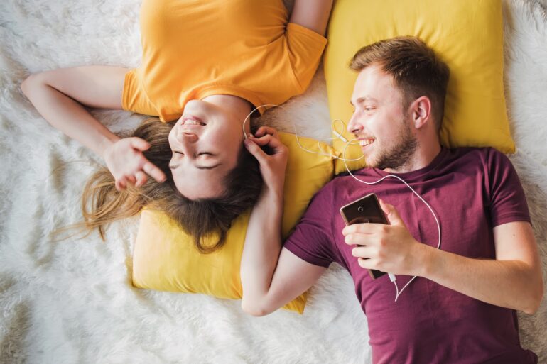 A man and woman on a blanket with pillows, sharing a pair of earbuds connected to a phone the man is holding.