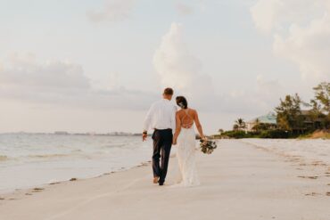 A bride and groom walk hand in hand along a beach coastline at sunset with soft waves and golden light.