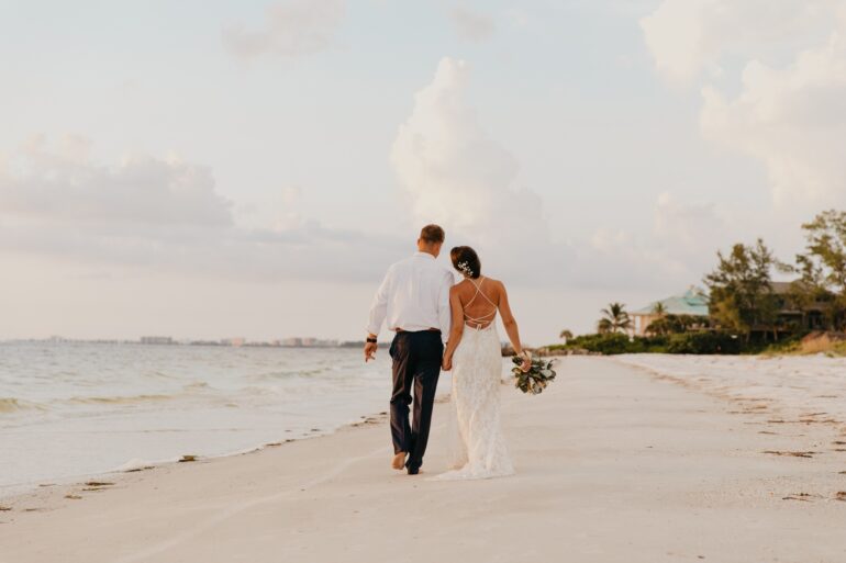 A bride and groom walk hand in hand along a beach coastline at sunset with soft waves and golden light.
