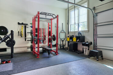 The interior of a residential garage setup as a gym with a free weight stand in the center of a soft, black mat.