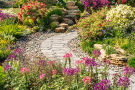 A stone path running through a small garden in a home’s yard. The garden is full of colorful flowers and green shrubs.